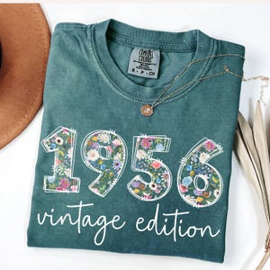 Könnte beinhalten: Ein teal Comfort Colors T-Shirt mit dem Jahr "1956" in floralem Design und den Worten "vintage edition" in weißer Schrift. Eine goldene Halskette mit einem kleinen Anhänger liegt auf dem Shirt.