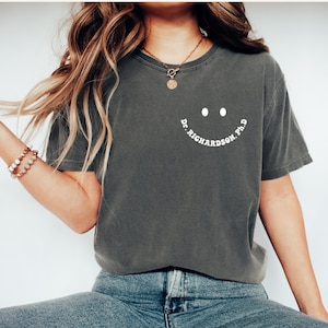 May include: A dark gray t-shirt with a white smiley face and the text "Dr. Richardson Ph.D." printed on the front.