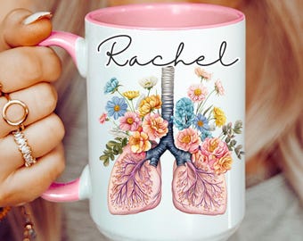 Puede incluir: Taza de cerámica blanca con borde y asa de color rosa. La taza presenta un diseño floral con un par de pulmones y el nombre "Rachel" en letra negra.