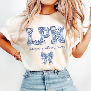 May include: Cream-colored t-shirt with the letters "LPN" in a blue floral design. Below the letters, the words "licensed practical nurse" are written in a cursive font. A blue floral bow is also featured on the shirt.