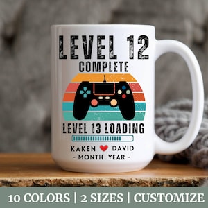 Könnte beinhalten: Weiße Keramiktasse mit einem Retro-Design, das einen Videospiel-Controller und den Text "Level 12 abgeschlossen, Level 13 wird geladen" zeigt. Die Tasse enthält auch eine Fortschrittsanzeige und den Text "Kaken David - Monat Jahr - ".