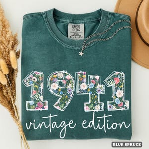 May include: Teal-coloured t-shirt featuring the year "1941" in a floral pattern, with the words "vintage edition". The shirt has a crew neck and short sleeves. A silver necklace with a star pendant is on the shirt.