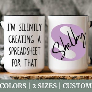 May include: Two white ceramic mugs with black handles. One mug has the text "I'm silently creating a spreadsheet for that." The other mug has a purple S with the name "Shelby" written on it.