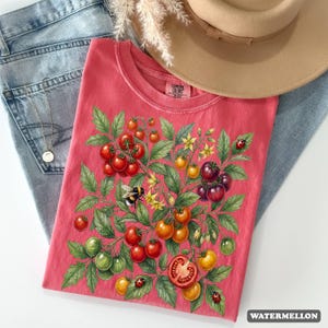 Op de afbeelding: Koraalrood T-shirt met een illustratie van een tomatenplant met rode, oranje en groene tomaten, gele bloemen, een hommel en lieveheersbeestjes. De tekst "WATERMELLON" staat rechtsonder.