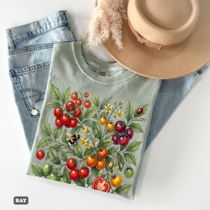 Op de afbeelding: Een lichtgroen T-shirt met een levendig tomatenplantontwerp met rode, oranje, gele en groene tomaten, een hommel en een lieveheersbeestje. Het shirt wordt gecombineerd met een lichtblauwe jeans en een beige hoed.