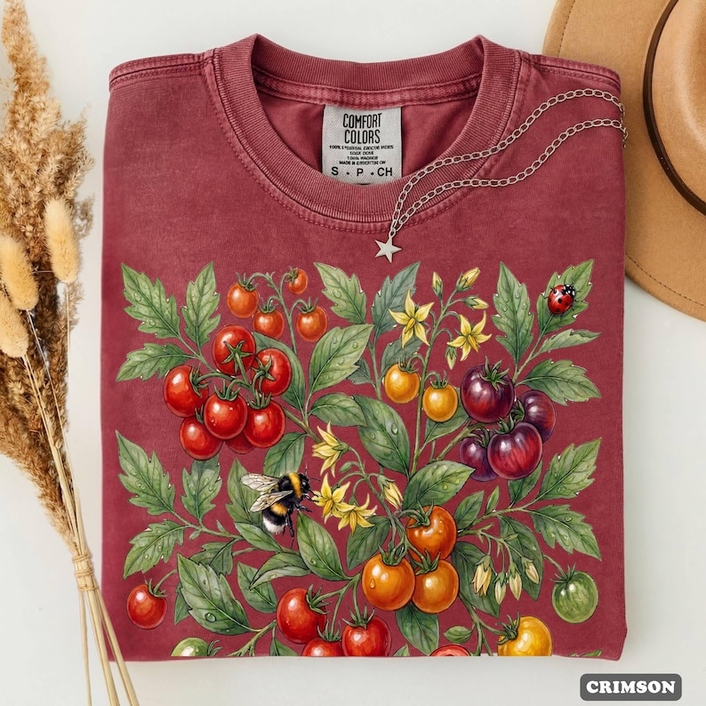 Op de afbeelding: Kleurrijk T-shirt met een illustratie van een tomatenplant. Het ontwerp bevat rode, oranje en paarse tomaten, groene bladeren, gele bloemen en een hommel. Een zilveren ketting en een Comfort Colors-label zijn zichtbaar.