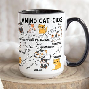 Może przedstawiać: Biały kubek ceramiczny z czarnym uchwytem. Kubek ma grafikę kota z wzorem chemicznym i tekstem "AMINO CAT-CIDS". Grafika zawiera tekst "LICK-SINE", "MEOWTAMIC ACID", "TRICKATPHAN", "PAWGININE", "HISSITIDINE", "CONTAMINE ASPURRTIC ACID" i "NEALANINE".