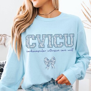 May include: Light blue sweatshirt with the text "CVICU" in a floral design. Below the text is the phrase "cardiovascular intensive care unit" in a script font. A floral bow graphic is centered below the text.