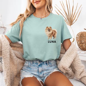 May include: A light blue t-shirt with a graphic of a small brown and white dog. The dog is standing on all four paws and looking to the right. The text "LUNA" is printed below the dog.