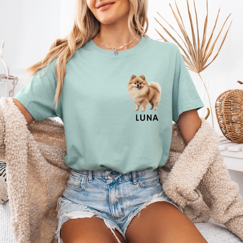 May include: A light blue t-shirt with a graphic of a small brown and white dog. The dog is standing on all four paws and looking to the right. The text "LUNA" is printed below the dog.