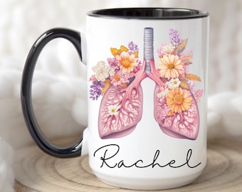 Puede incluir: Taza de cerámica blanca con borde y asa negros. La taza presenta una ilustración anatómica rosa y blanca de pulmones con flores que los rodean. El nombre "Rachel" está escrito en cursiva negra debajo de la ilustración.