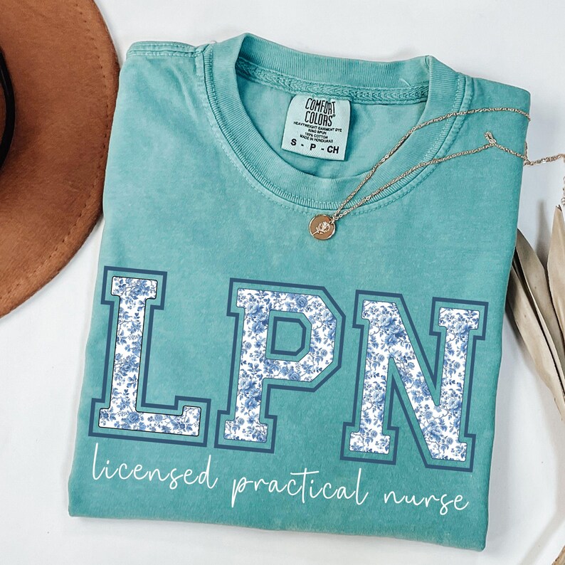 May include: Teal Comfort Colors t-shirt with the letters "LPN" in a blue floral pattern. The words "licensed practical nurse" are written below the letters in a script font. A gold necklace with a pendant is draped across the shirt.
