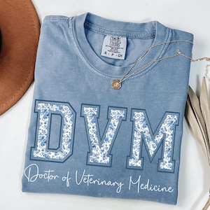 May include: A blue Comfort Colors t-shirt with the letters "DVM" in a floral pattern, and the words "Doctor of Veterinary Medicine" in a script font. A gold necklace with a pendant is draped across the shirt.