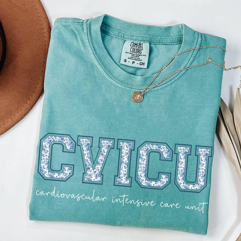 May include: A teal Comfort Colors t-shirt with the letters "CVICU" in a collegiate style, filled with a blue floral pattern. The words "cardiovascular intensive care unit" are printed below the letters. A gold necklace with a pendant rests on the shirt.