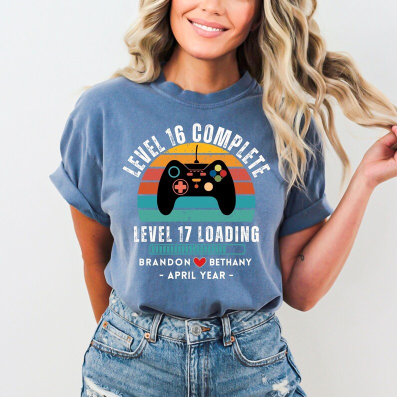 May include: A blue t-shirt with a retro-style video game controller graphic. The text on the shirt reads "Level 16 Complete Level 17 Loading Brandon &hearts; Bethany - April Year - "