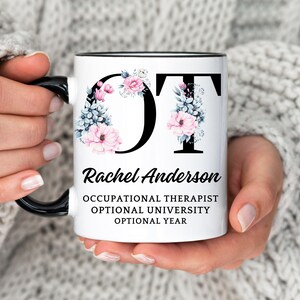 Puede incluir: Taza de cerámica blanca con borde y asa negros. La taza presenta un diseño floral con las letras "OT" en negro. El texto "Rachel Anderson Occupational Therapist Optional University Optional Year" está impreso debajo del diseño floral.