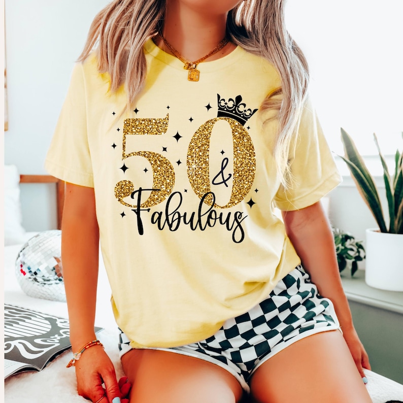 May include: A pale yellow short-sleeve t-shirt featuring a graphic print with the text 50 & Fabulous. The number 50 is rendered in a gold glitter texture, accompanied by a black crown icon, small black star accents, and black cursive lettering.
