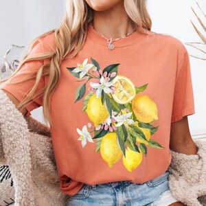 May include: Coral-colored t-shirt featuring a watercolor illustration of lemons, white blossoms, and green leaves. The design includes whole and sliced lemons, creating a vibrant, summery aesthetic. The shirt has a classic crew neck.