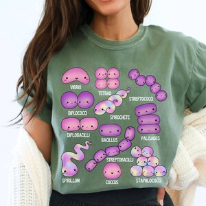 May include: Sage green t-shirt with a colourful illustration of different bacteria, such as Vibrio and Spirochete. Each bacterium is depicted with a cute, cartoon design and labelled with its scientific name. The t-shirt is made of a soft material.