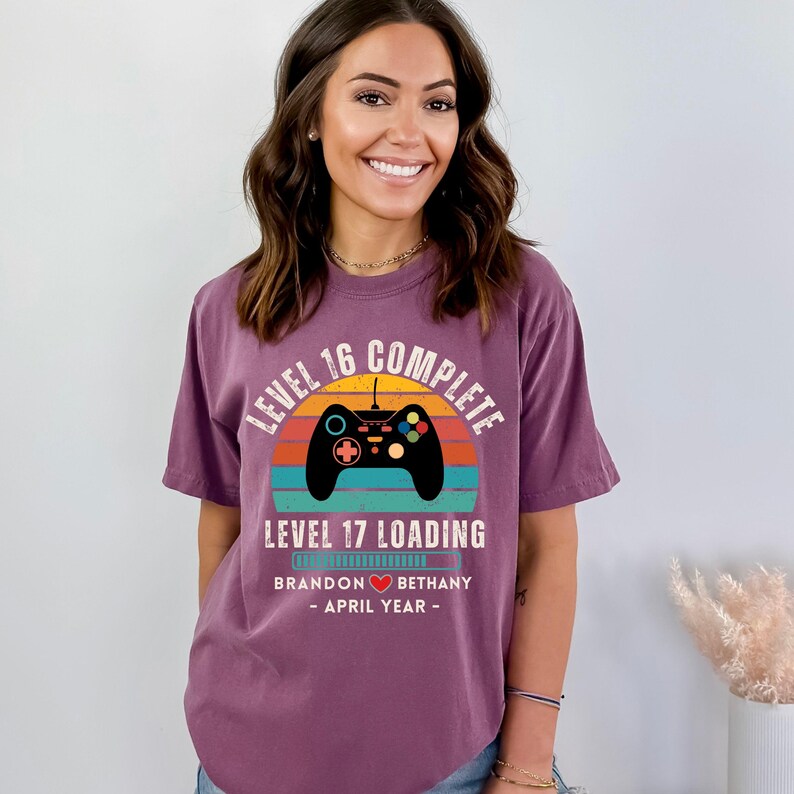 May include: Purple t-shirt with a retro-style graphic of a video game controller and the text "Level 16 Complete Level 17 Loading". The graphic also includes a sunset with a rainbow color scheme. The text "Brandon Bethany - April Year -" is below the graphic.