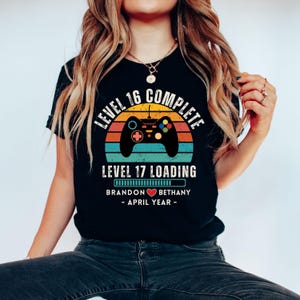 May include: Black t-shirt with a retro-style video game controller graphic. The text reads "Level 16 Complete" and "Level 17 Loading" with a progress bar. Below the progress bar is the text "Brandon &hearts; Bethany - April Year - "