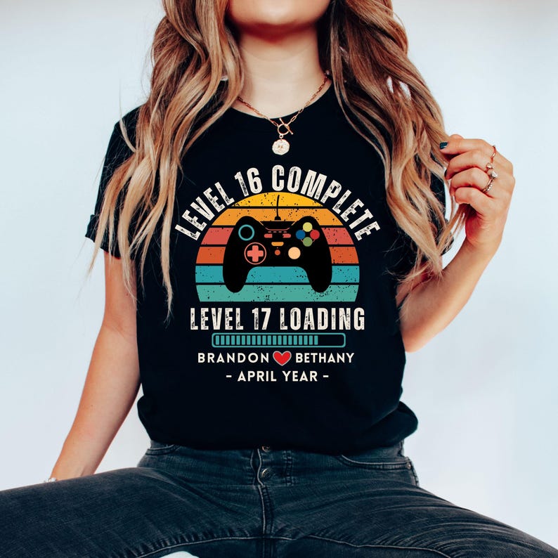 May include: Black t-shirt with a retro-style video game controller graphic. The text reads "Level 16 Complete" and "Level 17 Loading" with a progress bar. Below the progress bar is the text "Brandon &hearts; Bethany - April Year - "