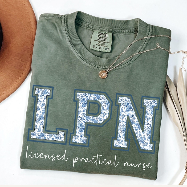 May include: An olive green t-shirt with the letters "LPN" in a blue floral pattern. The words "licensed practical nurse" are printed below the letters. A gold necklace and a brown hat are visible.