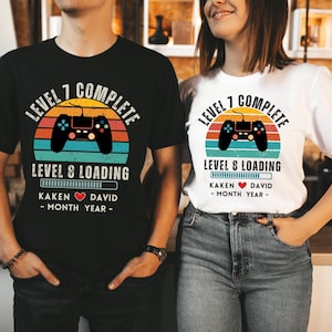 May include: Two black and white t-shirts with a retro video game controller graphic and the text "Level 7 Complete, Level 8 Loading". The shirts also include the names "Kaken" and "David" with a heart symbol between them and the text "Month Year".