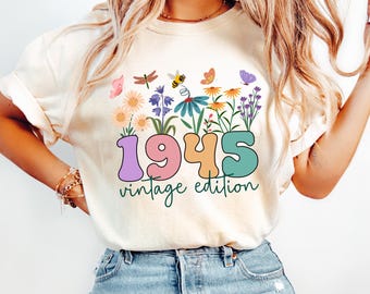 Può includere: T-shirt color crema con un design floreale colorato con farfalle, api e la scritta "1945 vintage edition" in un font retrò. Il design include fiori in vari colori, tra cui rosa, arancione e viola.