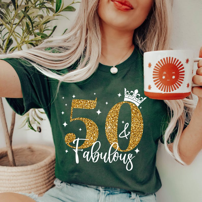 May include: A dark green t-shirt featuring a gold glitter graphic of the number 50 with a small crown on top and the text "& Fabulous" in white script. The shirt is paired with a white mug displaying a red sun illustration with a face.