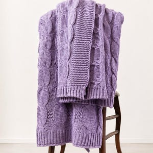 Peut inclure: Un plaid tricoté de couleur lavande présentant un motif de torsades proéminent combiné à des sections de texture côtelée. La couverture est drapée sur une chaise en bois, mettant en valeur la construction en fil épais et le travail des mailles.