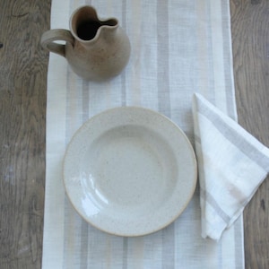 Peut inclure: Une cruche, une assiette et une serviette pliée en tons neutres sont disposées sur un chemin de table à rayures. La cruche a une anse et un bec incurvés. L'assiette est mouchetée et la serviette a des rayures grises. Sur une surface en bois.