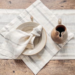 May include: A dining table arrangement with a beige plate, a knotted napkin, a small ceramic jug, and a striped linen placemat. The color scheme is composed of beige, grey, and brown hues, creating a warm and inviting atmosphere.
