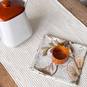 Peut inclure: Une théière en céramique blanche avec un couvercle marron est posée sur un chemin de table en lin rayé. Un petit bol orange et une cuillère en argent reposent sur une serviette à motifs floraux.