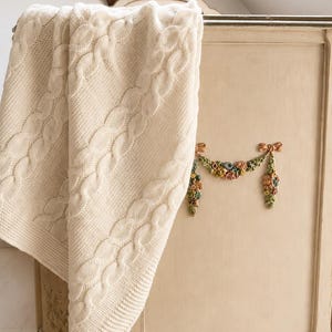 May include: A cream-colored cable knit blanket with a textured pattern draped over a light beige wooden cabinet. The cabinet features a decorative relief carving of a floral garland with small pink, blue, and green painted flowers and bows.