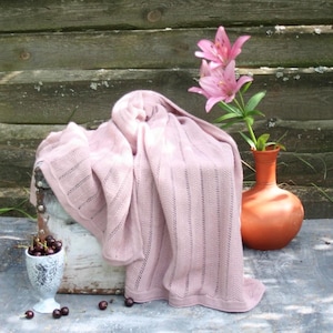 May include: A soft, light pink knitted blanket with a decorative edge is draped over a white chest. A terracotta vase holds pink lilies. A small, ornate cup is filled with cherries.