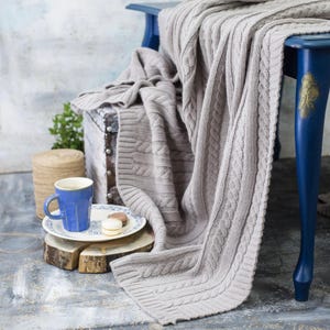 Peut inclure: Une couverture en tricot de couleur gris clair, avec un motif torsadé, drapée sur une chaise bleue et un coffre en bois. Une tasse bleue, une assiette avec deux macarons et une petite plante sont également visibles.