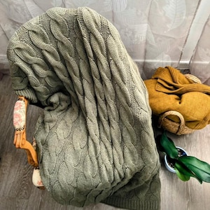 Peut inclure: Un plaid en tricot torsadé vert sauge drapé sur une chaise en bois. La couverture présente un motif tressé vertical proéminent. À côté de la chaise, une couverture en tricot jaune moutarde est rangée dans un panier en osier près d'une plante.