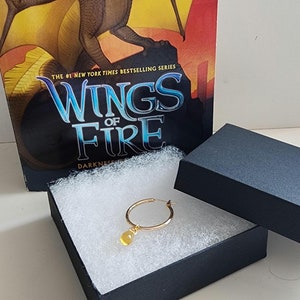 May include: A gold hoop earring with a yellow teardrop-shaped charm rests in a black box with white filler, next to a book titled "Wings of Fire: Darkness of Dragons". The book cover features a dragon.