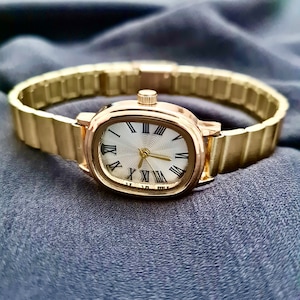 May include: A gold-tone wristwatch with a white face and black Roman numeral hour markers. The watch has a gold-tone metal band.