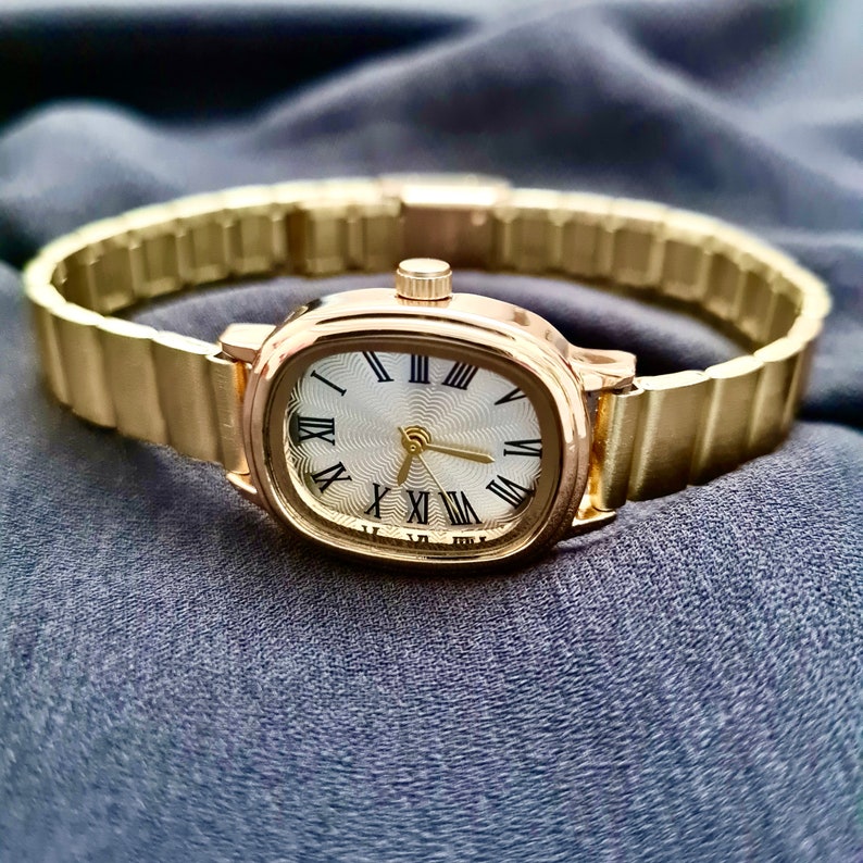 May include: A gold-tone wristwatch with a white face and black Roman numeral hour markers. The watch has a gold-tone metal band.
