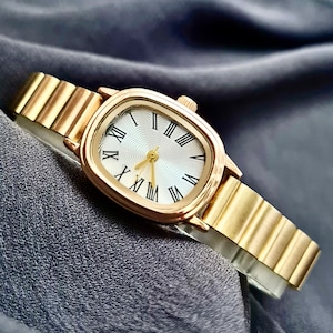 May include: A gold-tone wristwatch with a square face and Roman numeral hour markers. The watch has a gold-tone metal band.