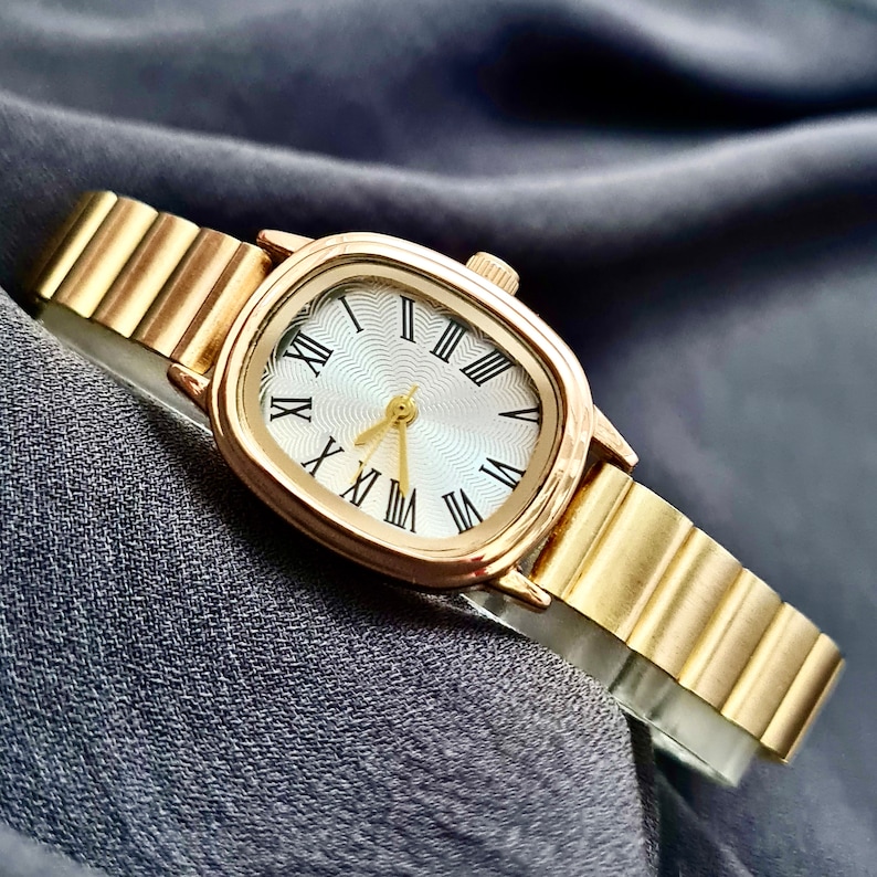 May include: A gold-tone wristwatch with a square face and Roman numeral hour markers. The watch has a gold-tone metal band.
