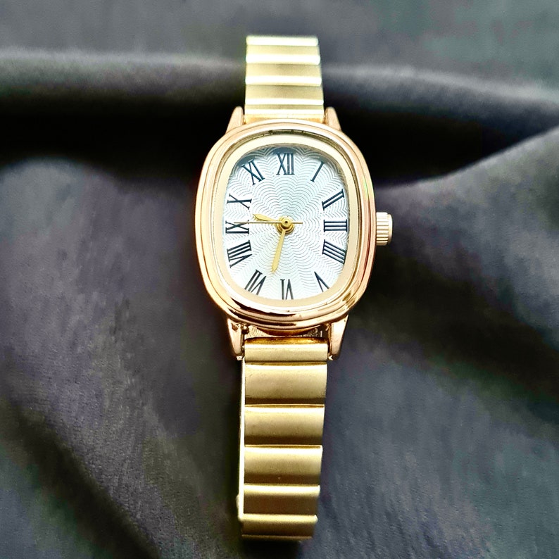 May include: A gold-tone wristwatch with a white face and Roman numeral hour markers. The watch has a square-shaped face and a metal band.