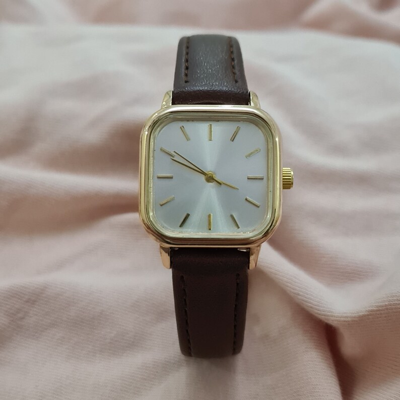Puede incluir: Un reloj de pulsera de cara cuadrada con una correa de cuero marr&oacute;n y una caja de tono dorado. La esfera del reloj es blanca con manecillas y marcadores de hora de tono dorado.