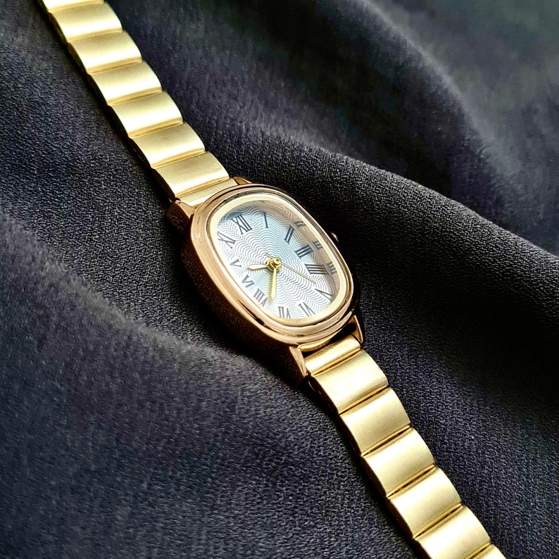 May include: A gold-tone watch with a rectangular face and Roman numeral hour markers. The watch has a gold-tone metal band.