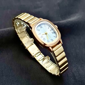 May include: A gold-toned wristwatch with a square face and a blue dial. The watch has Roman numeral hour markers and a gold-toned metal band.