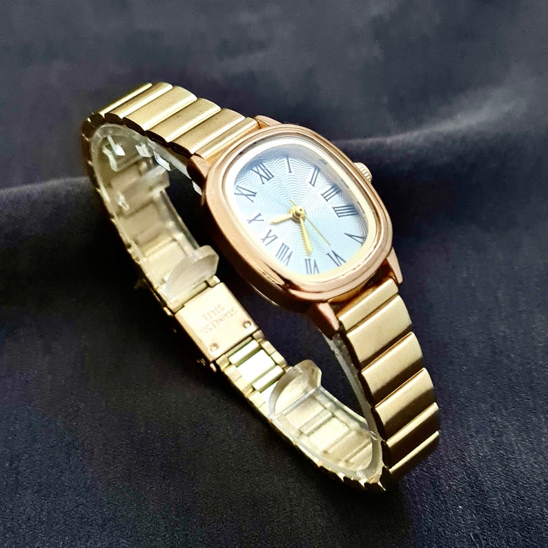 May include: A gold-toned wristwatch with a square face and a blue dial. The watch has Roman numeral hour markers and a gold-toned metal band.