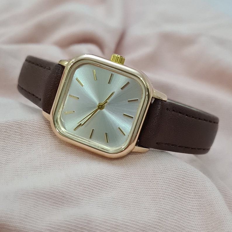 Puede incluir: Un reloj de pulsera de forma cuadrada con una caja de color dorado y una correa de cuero marr&oacute;n. La esfera del reloj es blanca con agujas e &iacute;ndices de color dorado.