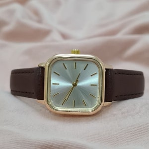 Puede incluir: Un reloj de pulsera de forma cuadrada con una correa de cuero marr&oacute;n y una caja dorada. La esfera del reloj es plateada con agujas e &iacute;ndices dorados.